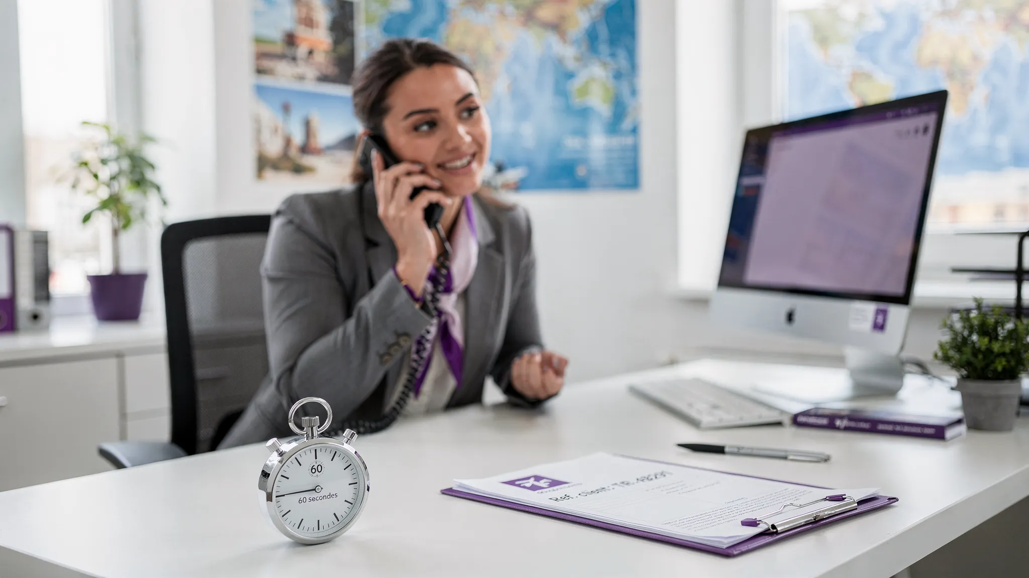 Une conseillère d’agence de voyage explique au téléphone un paiement par virement instantané, avec un petit chronomètre “60 secondes” posé sur le bureau, et un dossier client papier avec une référence visible.